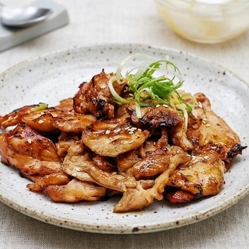 Grilled chicken pieces with a glossy glaze, sesame seeds, and green onions on a speckled plate.