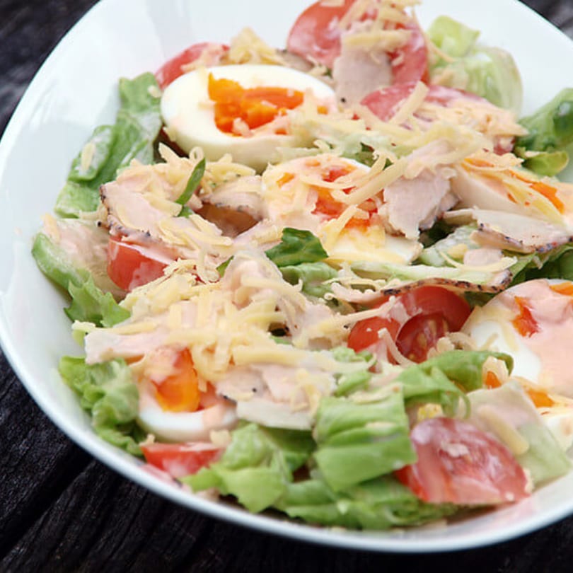 A fresh salad in a white bowl, featuring lettuce, tomatoes, sliced eggs, and shredded cheese.