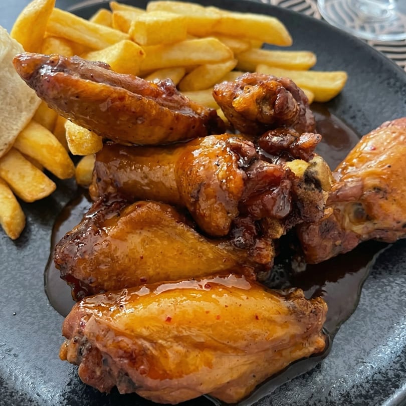 Glazed chicken wings and golden french fries on a dark plate, with a glass in the background.
