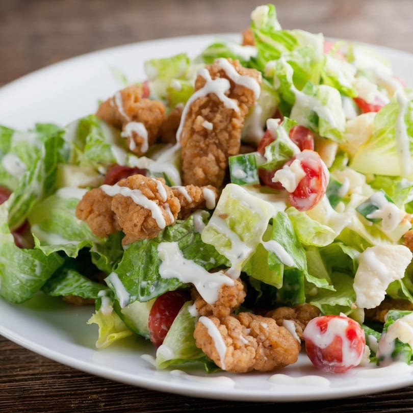 Crisp green salad with fried chicken pieces, cherry tomatoes, and creamy dressing on a white plate.